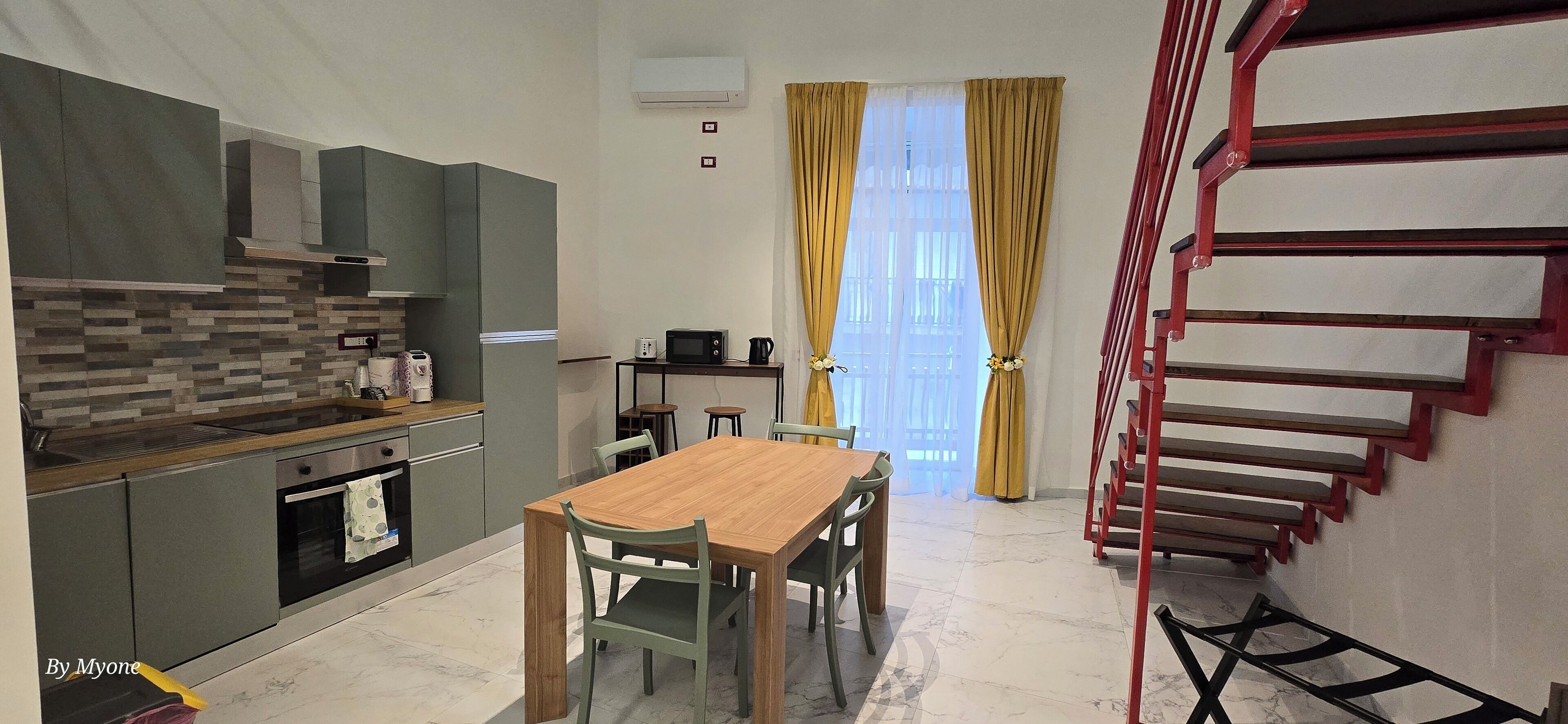 A modern kitchen with grey cabinets, a wooden countertop, and a built-in oven. There is a dining area with a wooden table and chairs. A staircase with red railings leads to an upper level.