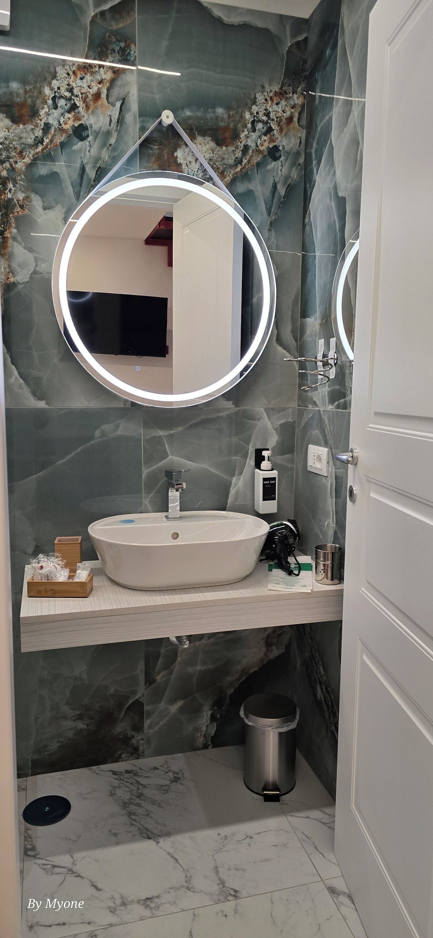 A bathroom with a marble sink, two mirrors, a handheld showerhead, and a trash bin.