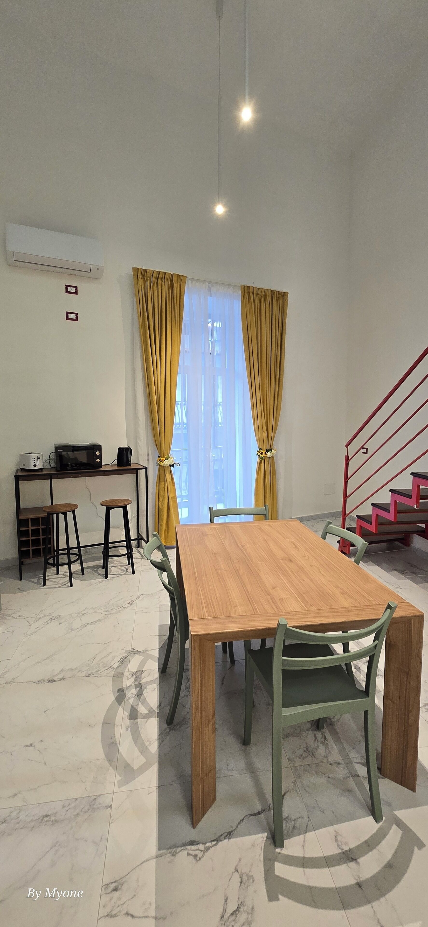 A dining area with a wooden table and chairs, a kitchen area with a microwave and toaster, and a staircase with a red railing.