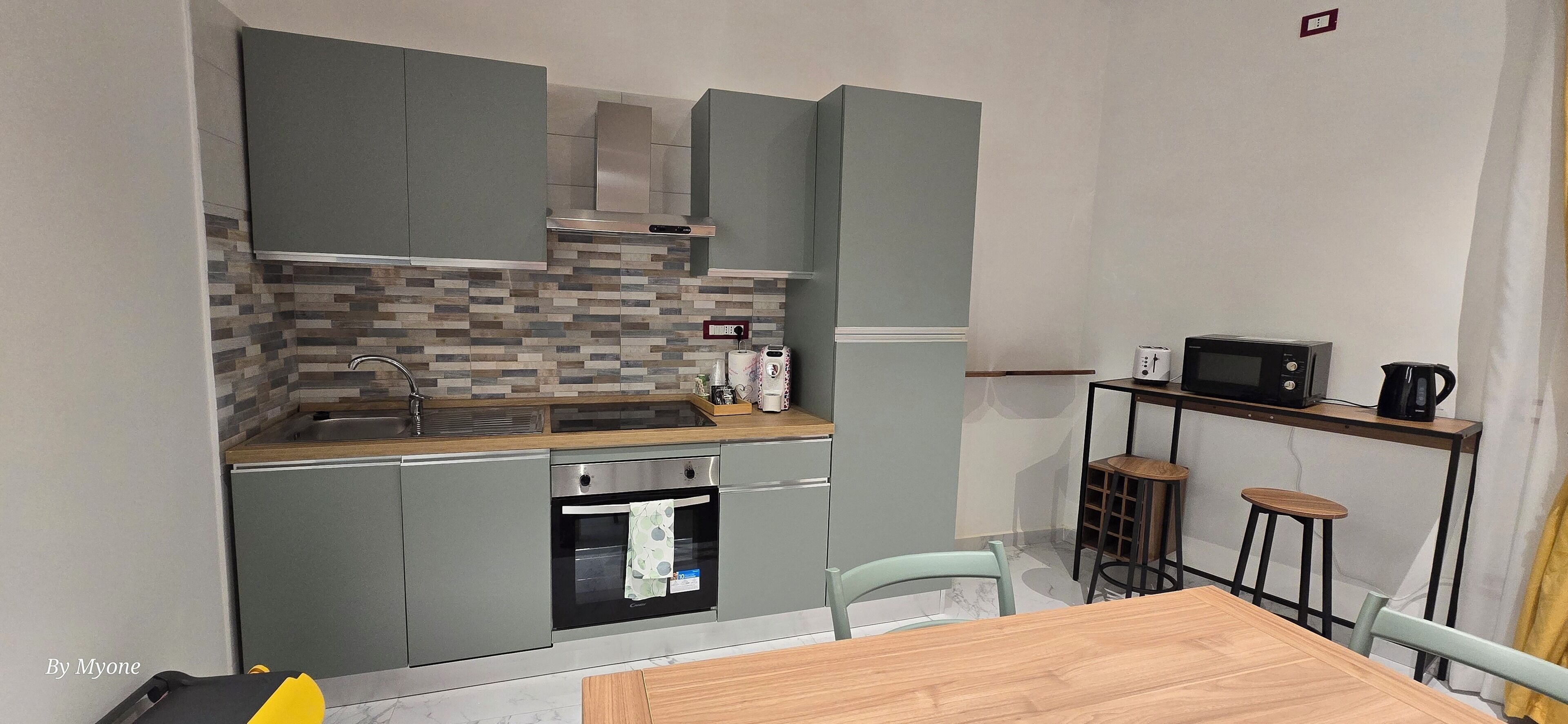 A compact kitchen with grey cabinets, a tiled backsplash, a built-in oven, a microwave, and a small dining area with stools.