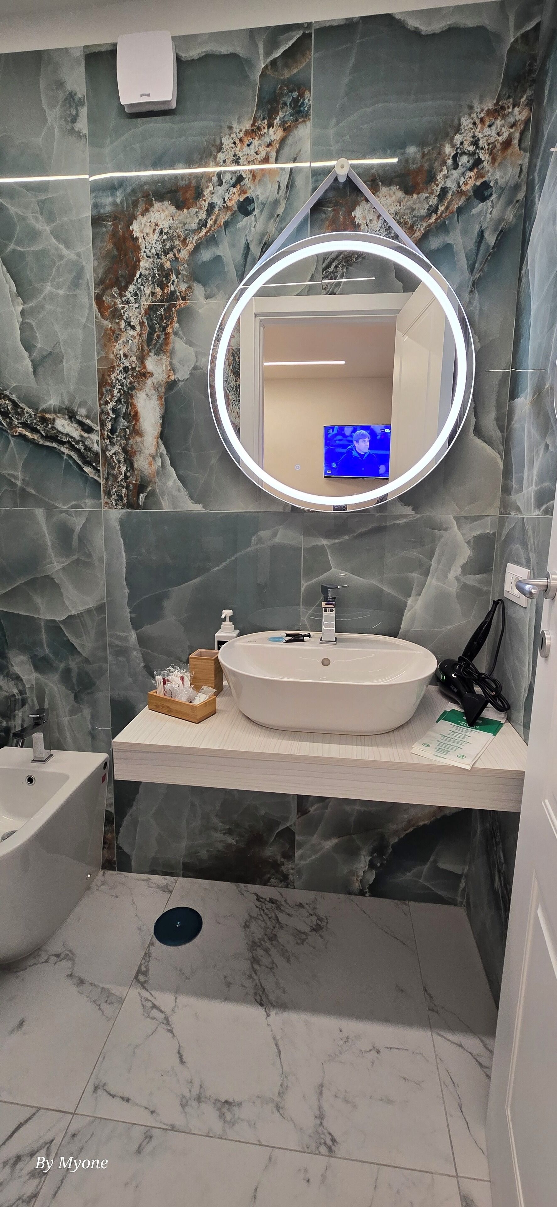 A bathroom with a marble wall, a round mirror, a sink, and a handheld showerhead.