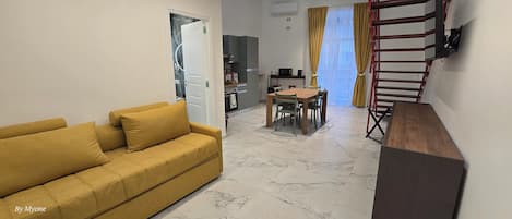 A living room with a yellow sofa, a dining table with chairs, a kitchen area, and a staircase.