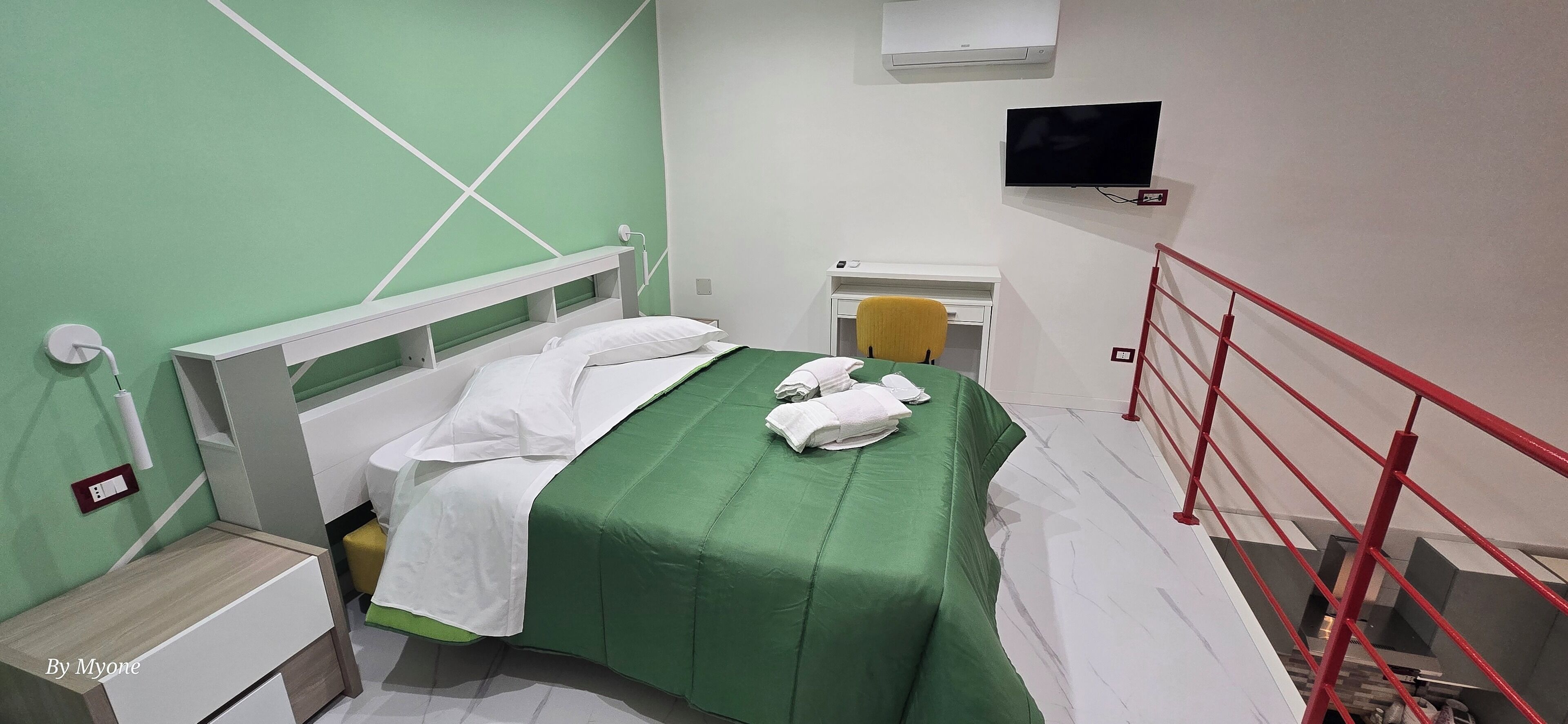 Ein Hotelzimmer mit einem Bett, einem Schreibtisch, einem Sessel und einem Fernseher, sowie einer roten Geländer.