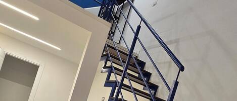 Ein modernes Interieur mit Blau- und Weißtönen, mit einer Treppe aus dunklem Holz und blauen Geländern, einem türkisfarbenen Sofa und einem Marmorboden.