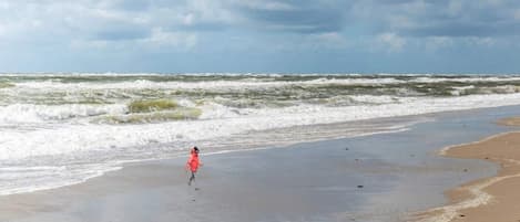 A beach with waves and a person in a red jacket running towards the water.