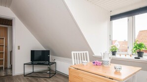 A room with a sloped ceiling, a TV on a black stand, a wooden desk, a white chair, a radiator, and a window with potted plants.