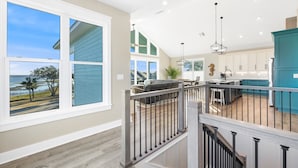 A spacious living area with a large window overlooking a beach, a kitchen with teal cabinets, and a staircase leading to an upper level. - Amazing Gulf views ~ 4 bedrooms ~ Brand new home ~ Great for families (Cedar Key)