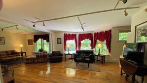 Amplia sala de estar con piso de madera, un sofá, dos sillones, una mesa de centro y un televisor. Ventanas grandes con cortinas rojas ofrecen luz natural y vistas a zonas verdes.
