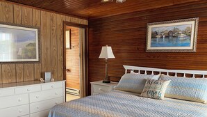 Un dormitorio con pared de paneles de madera, una cama con colcha a rayas, una mesita de noche con lámpara y un cuadro enmarcado sobre la cama.