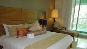 A hotel room with a large bed, a bedside table with a lamp, a chair, and a view of greenery through the window.