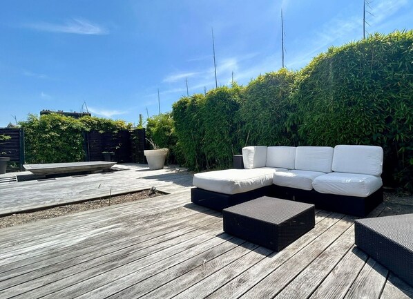 A rooftop patio with a white sectional sofa, a black rectangular ottoman, and a wooden deck.