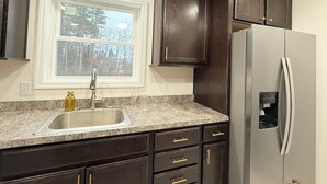 A kitchen with dark wood cabinets, a stainless steel refrigerator, a granite countertop, and a window with a view of trees.