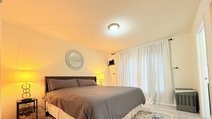 A bedroom with a bed, two bedside lamps, a wall mirror, a white curtain, a wicker basket, and a marble-patterned rug.