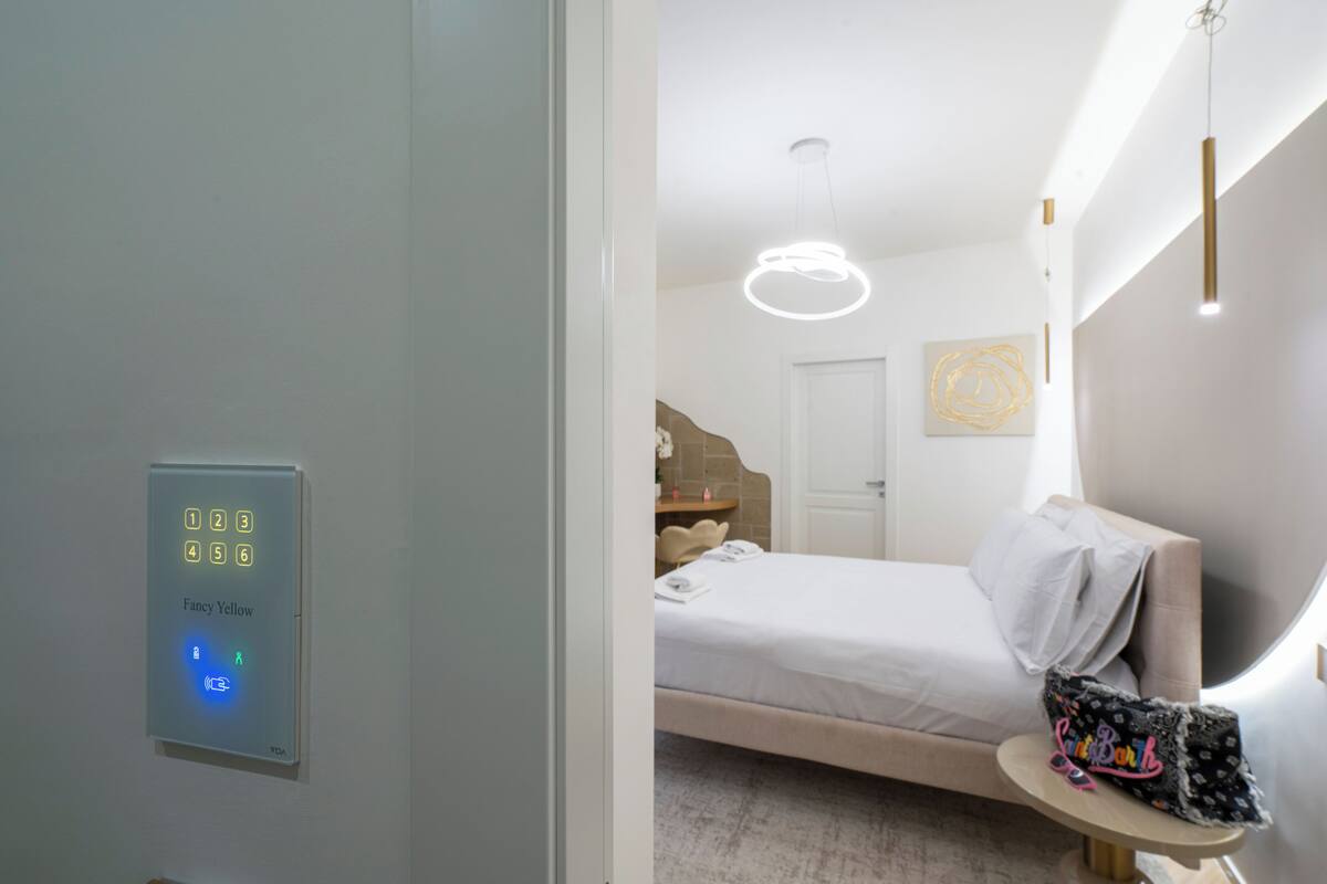 A modern hotel room with a bed, bedside table, and a wall-mounted control panel.