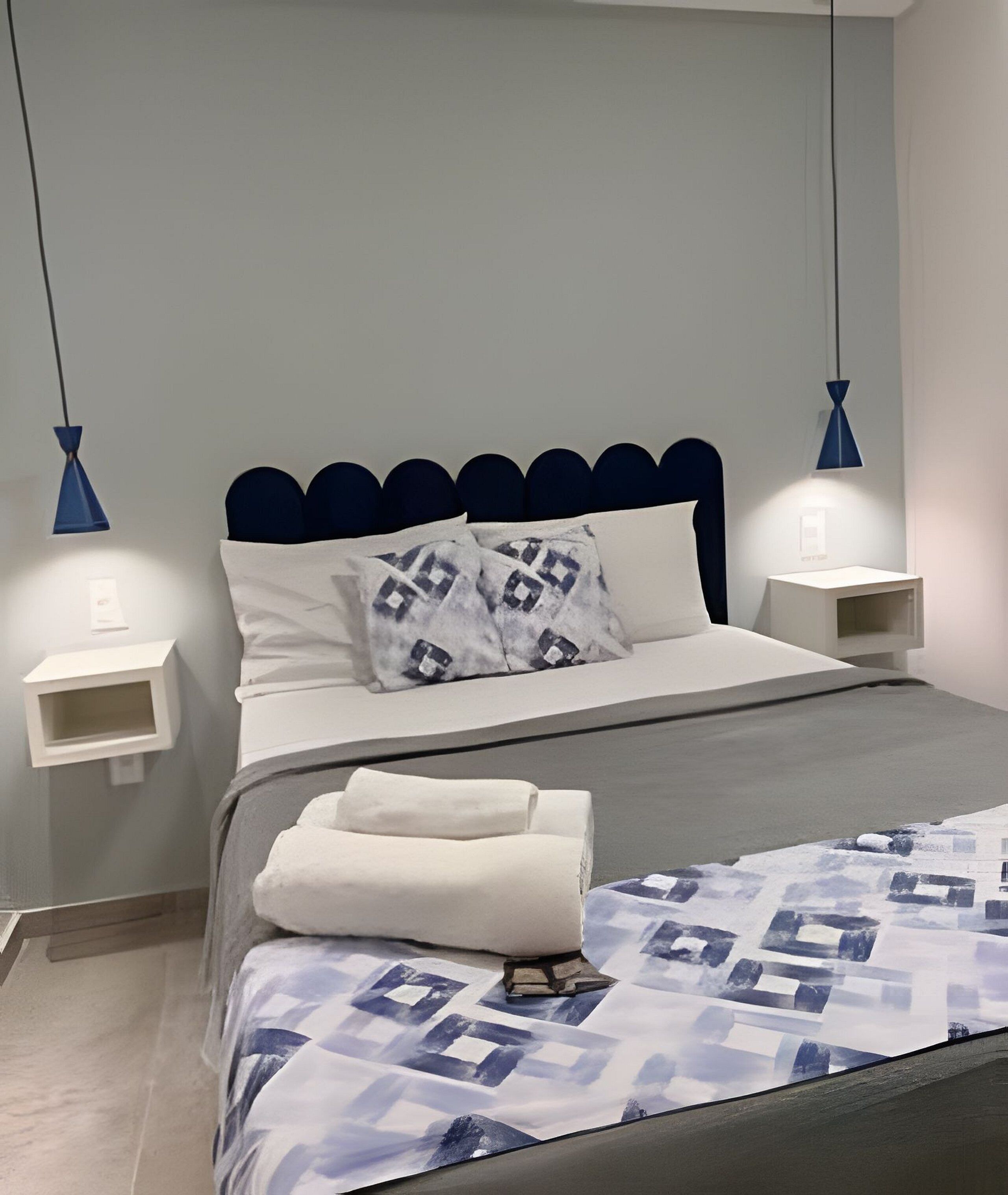 A neatly made bed with a patterned bedspread, two pillows, and a folded towel. There are two bedside tables with lamps, and the room has a modern design with a blue accent wall.