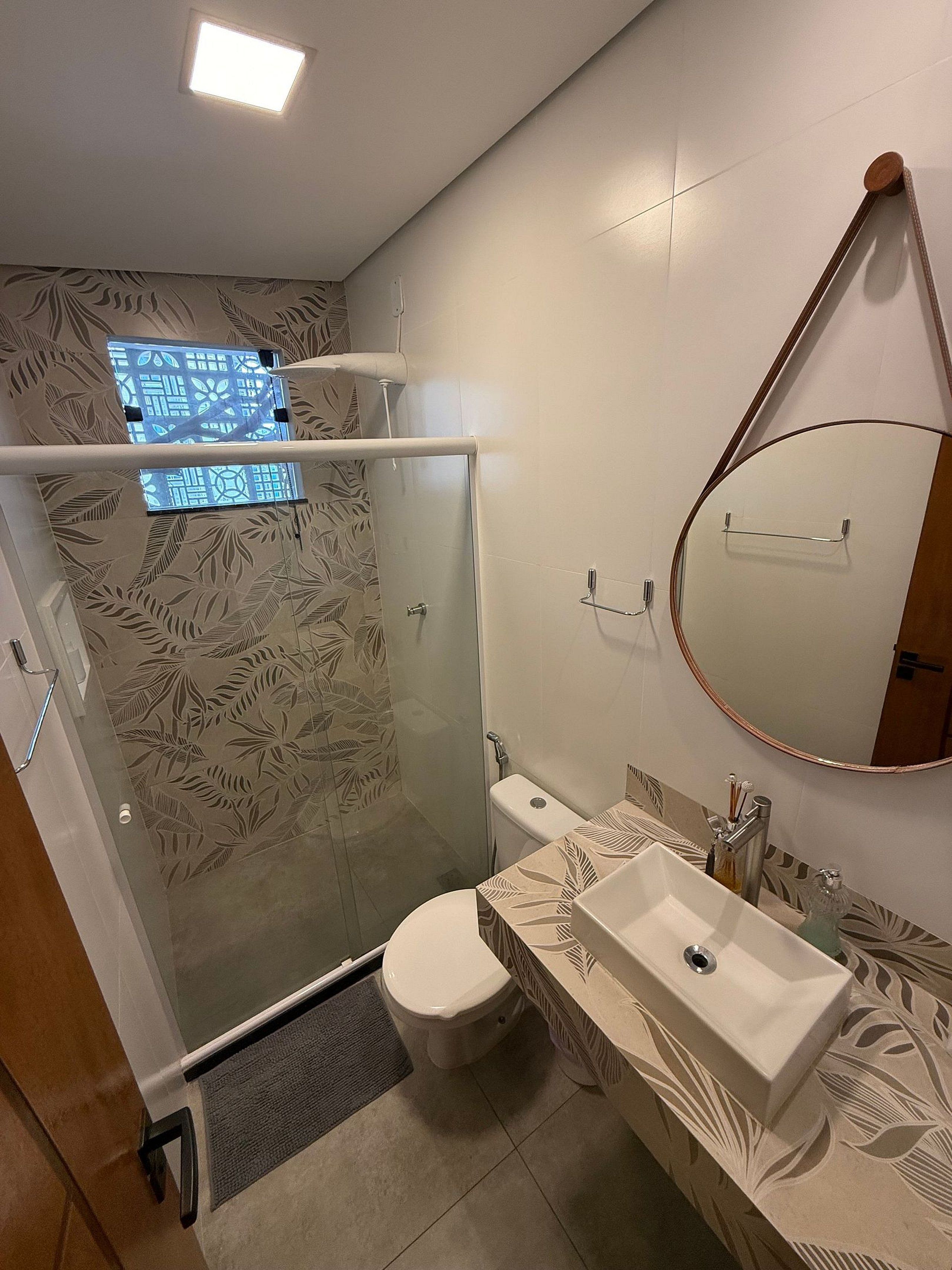 Casa de banho com cabine de duche em vidro, sanita, lavatório com espelho e azulejos estampados.