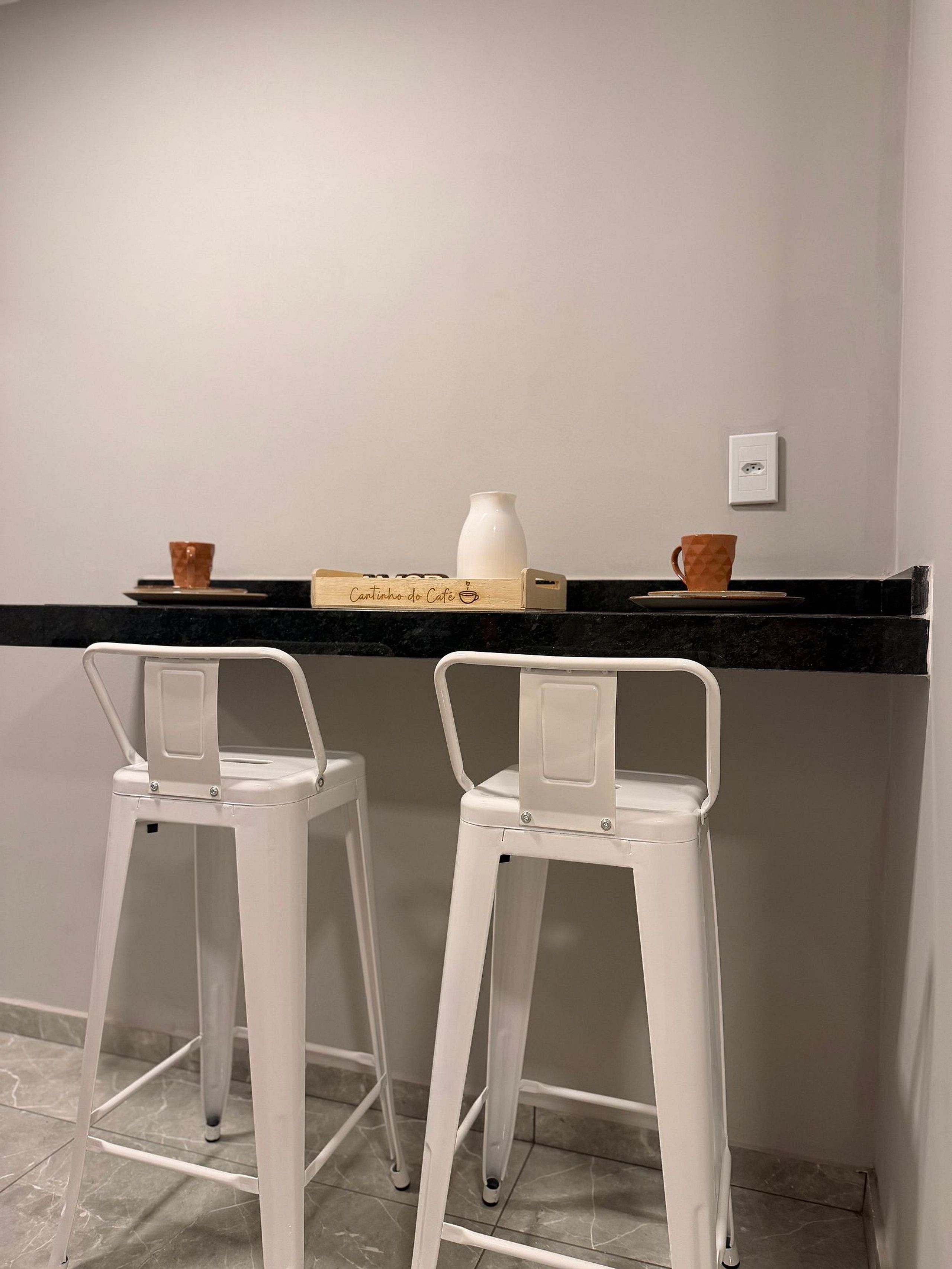 A kitchen area with a black countertop, two white bar stools, a white vase, and a wooden box labeled 'Cantinho do Café'.