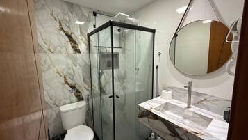 A bathroom with a glass-enclosed shower, a toilet, a sink with a marble countertop, and a round mirror.