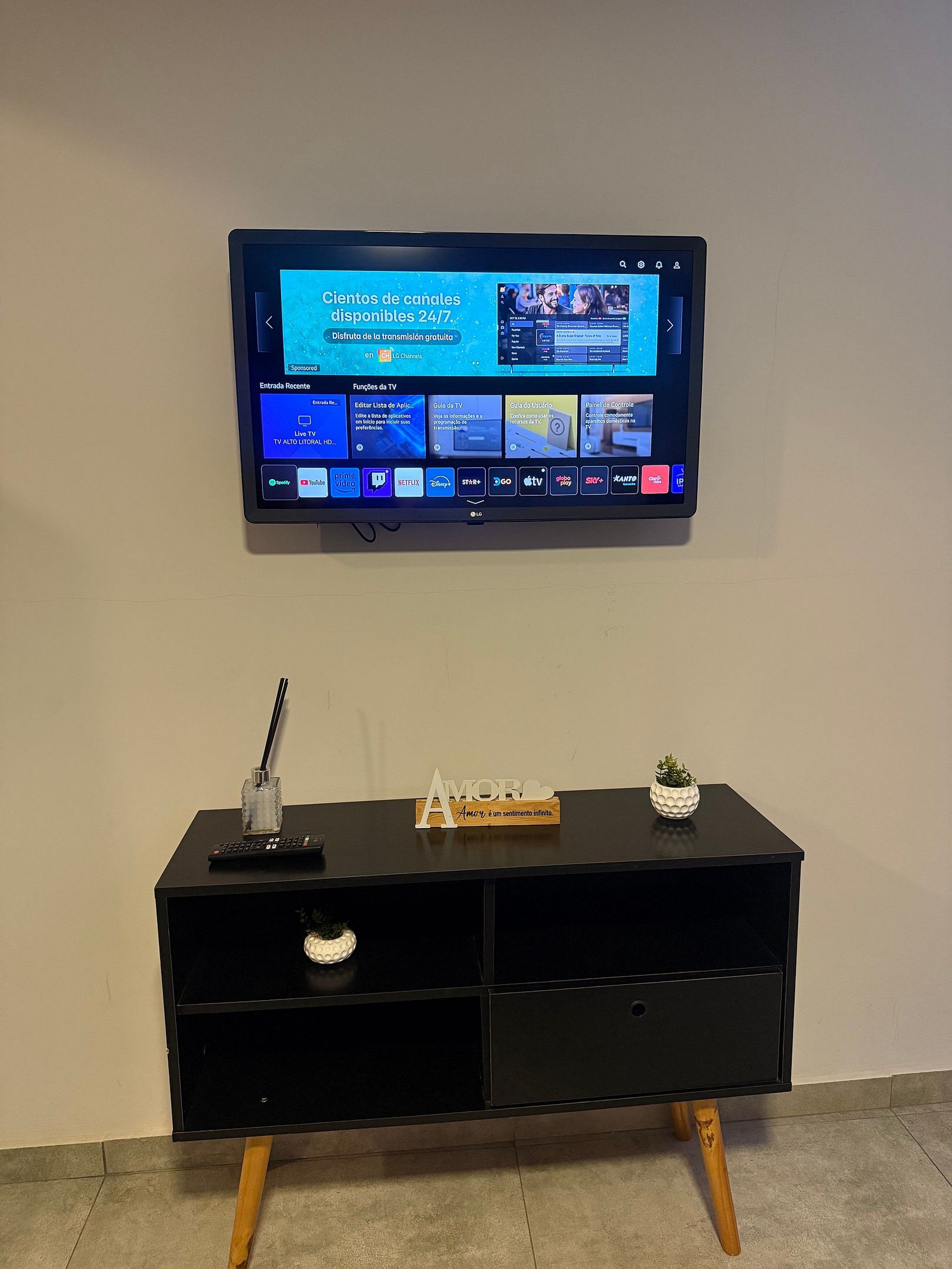Un televisor de pantalla plana montado en la pared, sobre un mueble negro para televisión, con un control remoto y objetos decorativos.