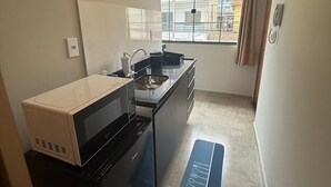 Een kleine keuken met een magnetron, kookplaat en spoelbak, een raam met gordijnen, en een blauwe mat met keukengerei.