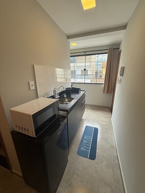 Una cocina compacta con microondas, estufa y lavaplatos, una ventana con cortinas, y un tapete azul con utensilios de cocina.