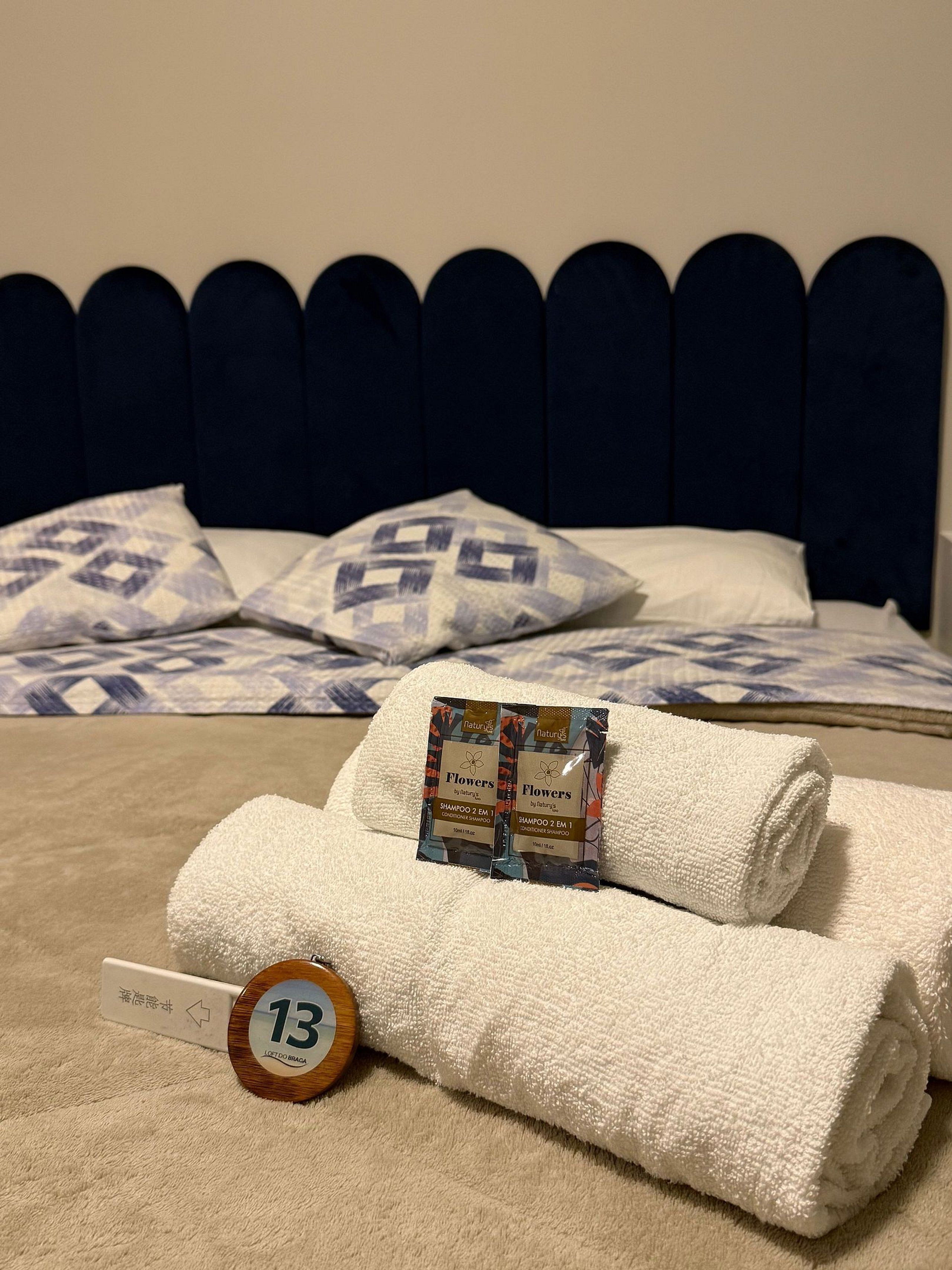 Una cama bien hecha con cabecera oscura, dos almohadas estampadas y una toalla blanca enrollada con un paquete de champú.