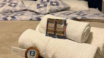 Una cama bien hecha con cabecera oscura, dos almohadas estampadas y una toalla blanca enrollada con un paquete de champú.