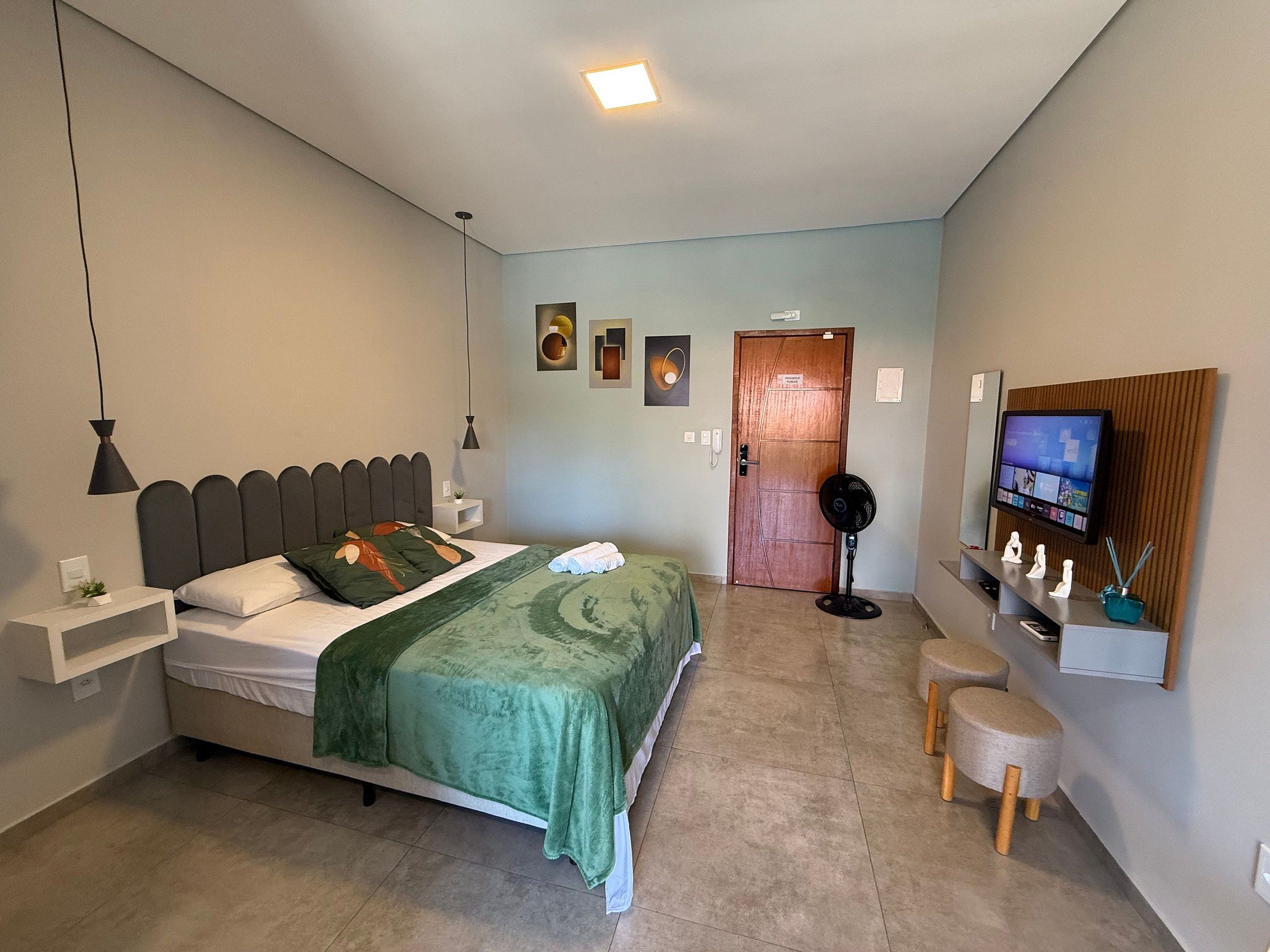 Un dormitorio con cama, mesitas de noche, televisor, ventilador y una puerta.