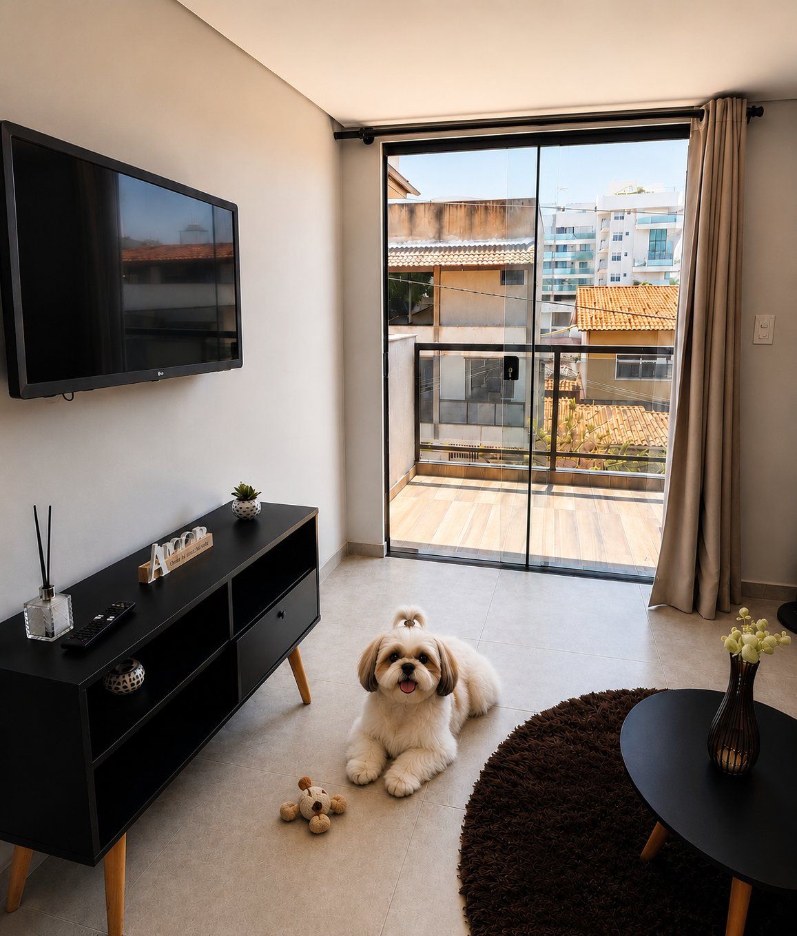 Un salón moderno con televisor, mueble de entretenimiento negro, un perro y vista a un barrio residencial a través de una puerta corrediza de vidrio.