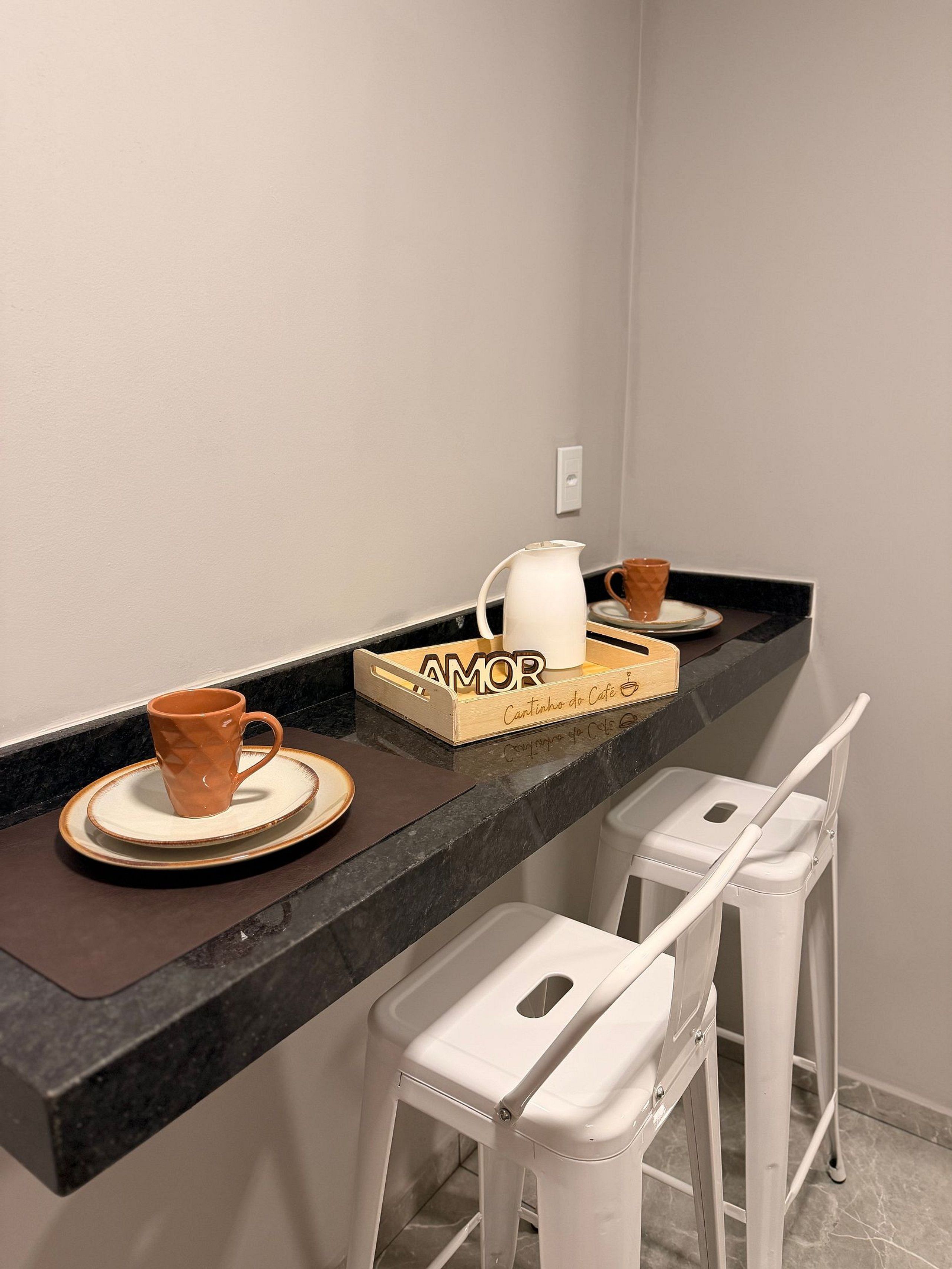 Un piano di lavoro in cucina con una macchina per il caffè, una tazza e una scatola con la scritta 'AMOR'. Ci sono due sgabelli da bar bianchi e un cestino della spazzatura sotto.