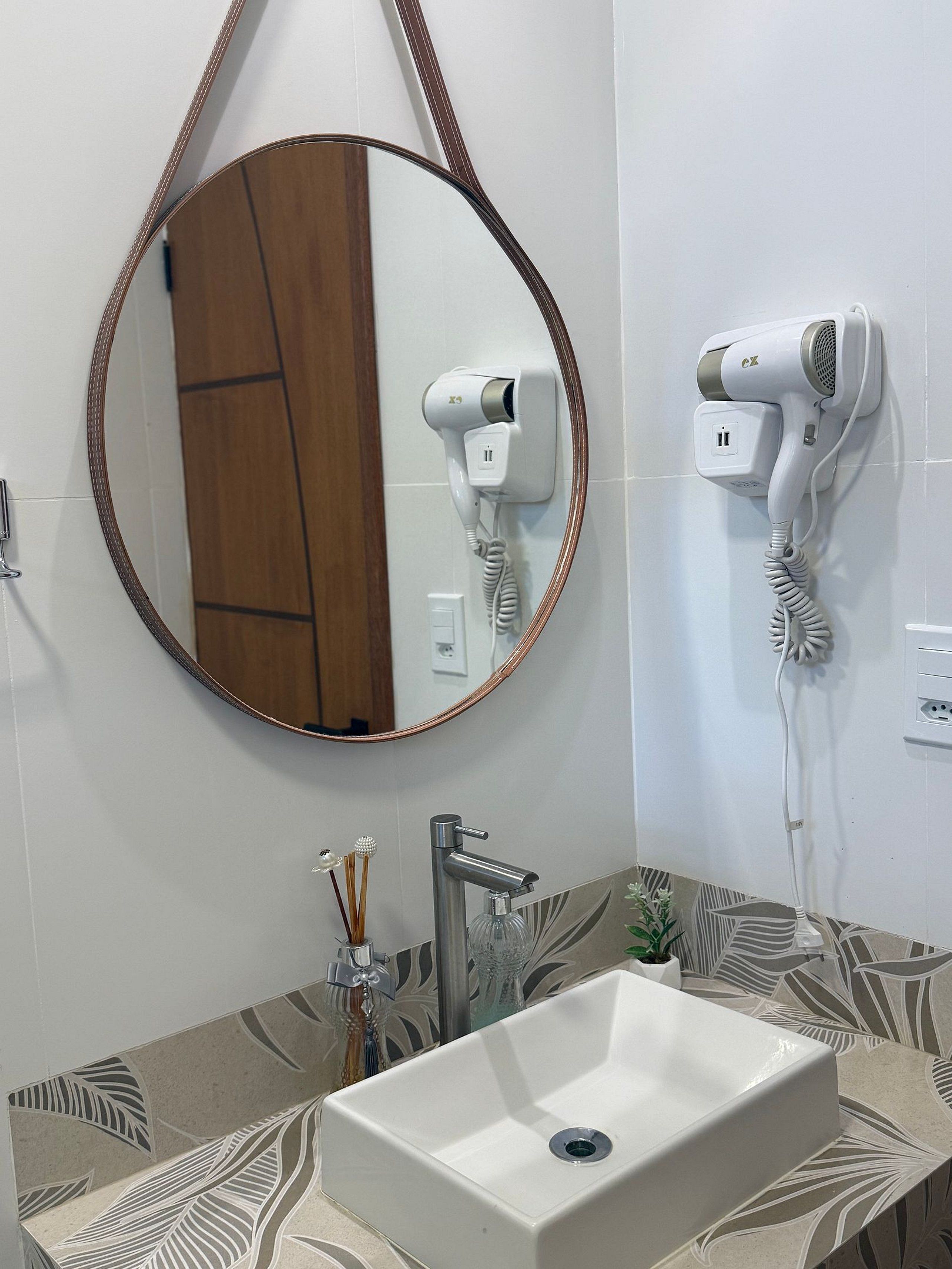 Ein Badezimmer mit einem rechteckigen Waschbecken, einem runden Spiegel und zwei an der Wand befestigten Haartrocknern.