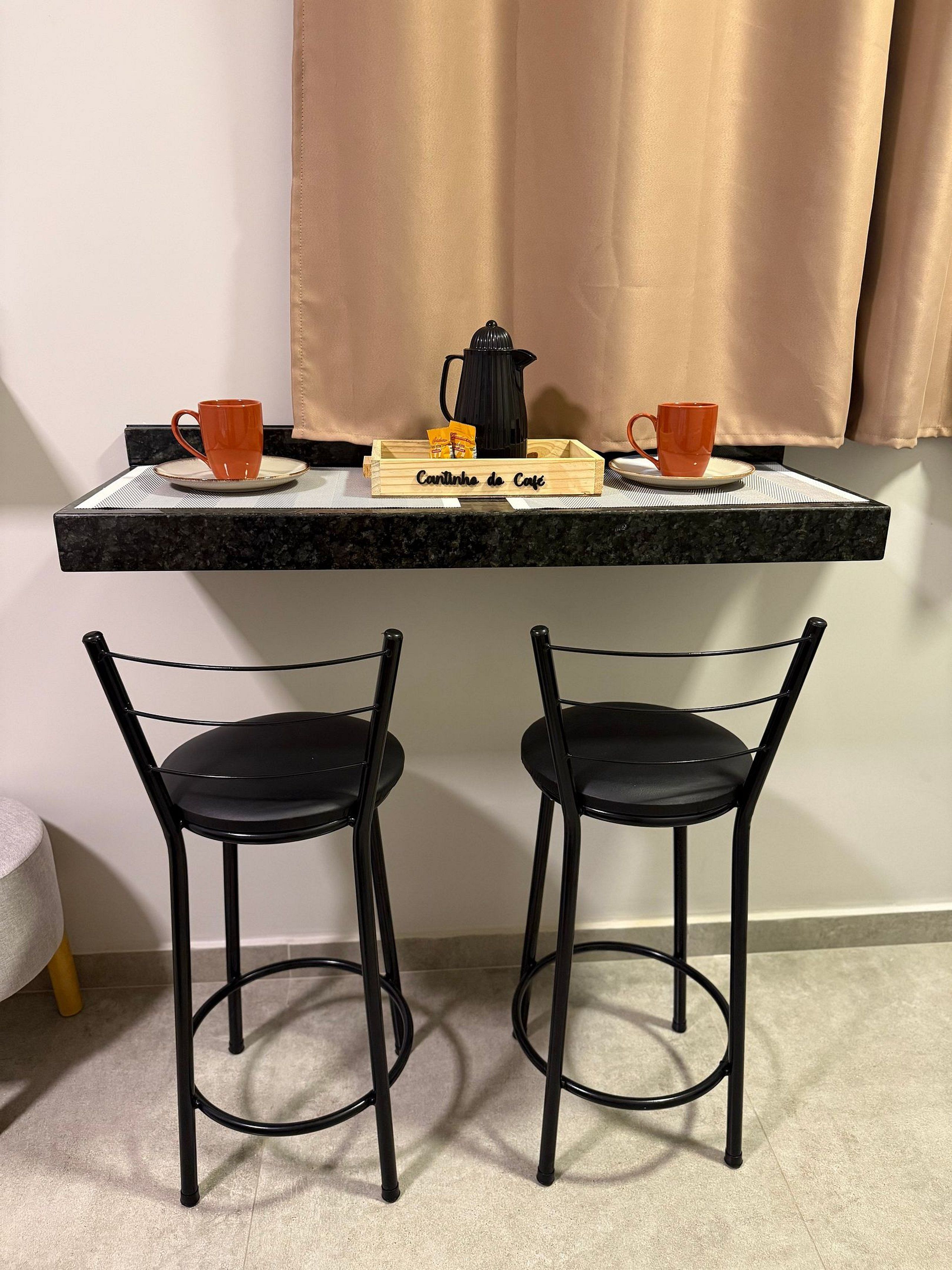 Uma pequena área de cozinha com uma bandeja de madeira identificada como 'Cantinho de Café', contendo duas xícaras laranjas, uma cafeteira preta e uma embalagem de café. Abaixo, duas cadeiras de metal pretas.