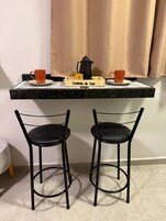 Una pequeña cocina con una bandeja de madera con la etiqueta 'Cantinho de Café' que contiene dos tazas naranjas, una cafetera negra y un paquete de café. Abajo, hay dos sillas de metal negras.