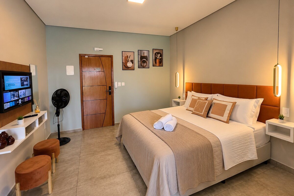 A hotel room with a bed, bedside tables, a TV, a fan, and a desk with a chair.