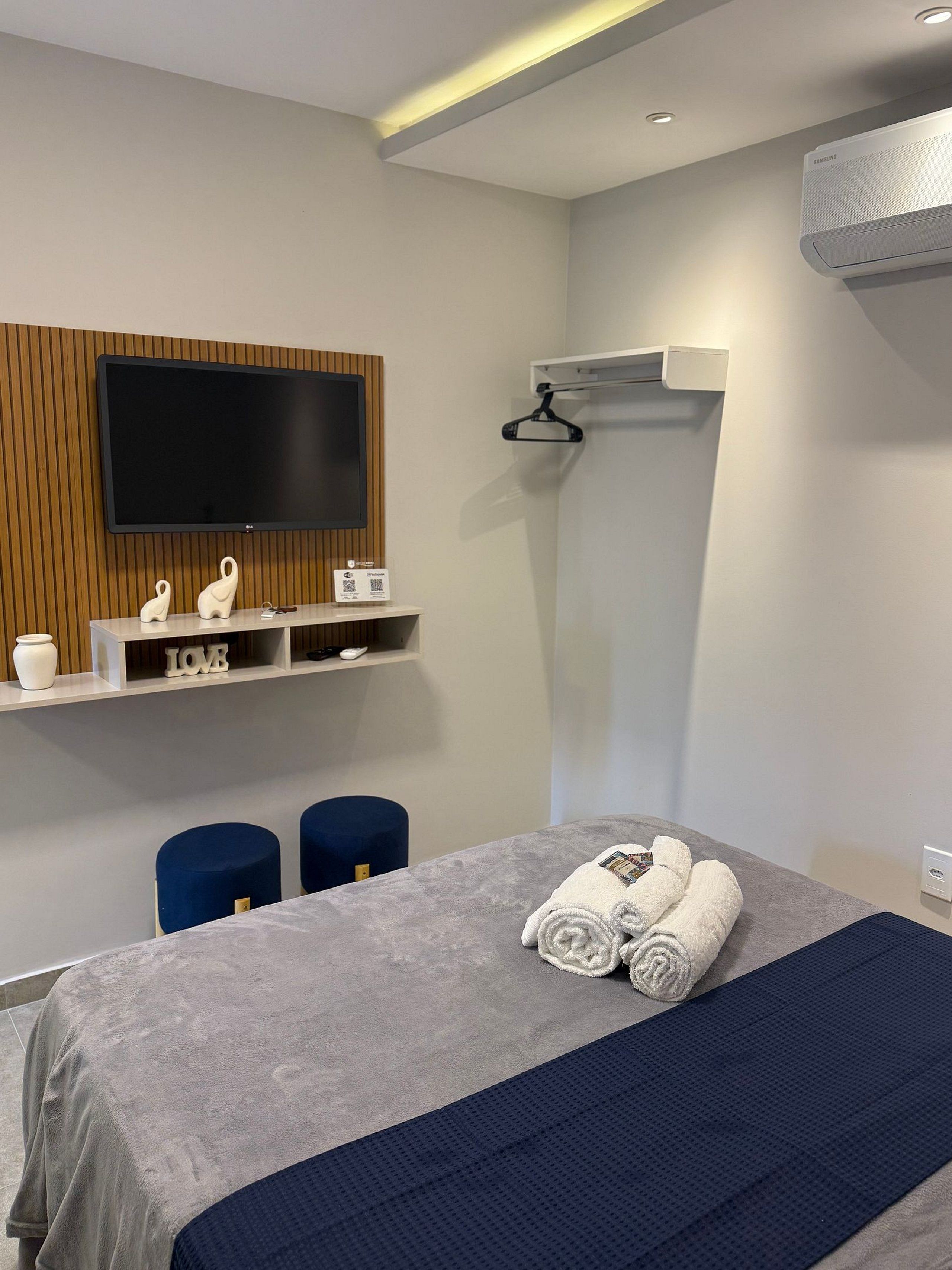 Ein modernes Hotelzimmer mit Flachbildfernseher, Wandklimaanlage, einem Bett mit grauer Decke und gerollten Handtüchern sowie einem kleinen Regal mit Dekoration.