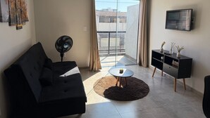Een moderne woonkamer met een zwarte bank, een ronde salontafel, een ventilator, een televisie en een balkon met uitzicht op gebouwen.
