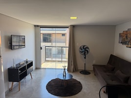 A living room with a sofa, a TV mounted on the wall, a standing fan, a small table with a vase, and a sliding glass door leading to an outdoor area.