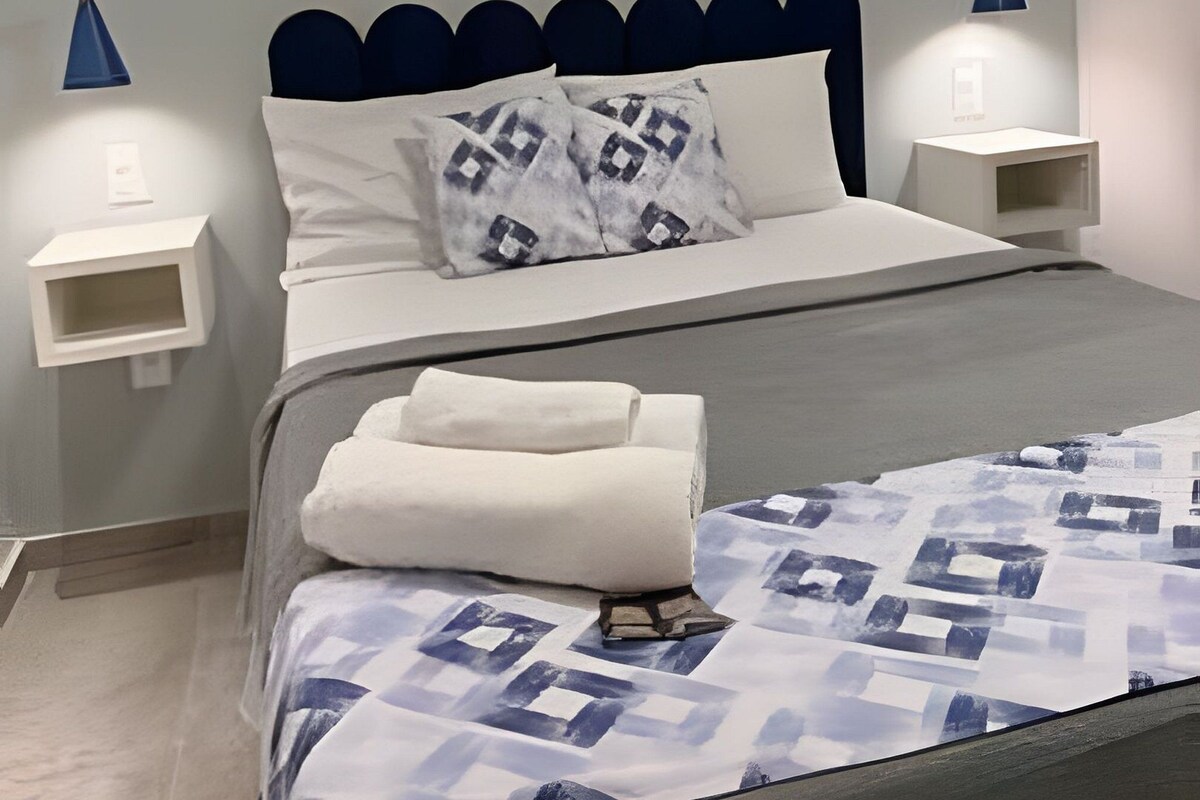A neatly made bed with a patterned bedspread, two pillows, and a folded towel. There are two bedside tables with lamps, and the room has a modern design with a blue accent wall.