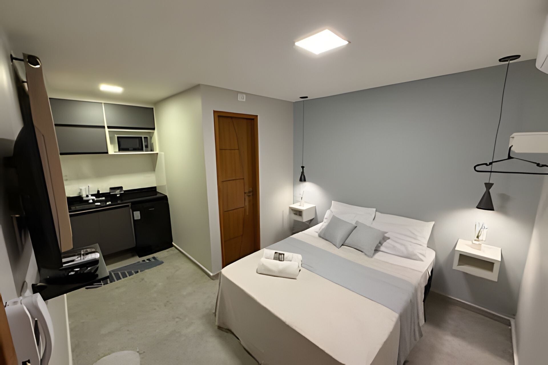 A hotel room with a bed, bedside table, lamp, and a view of the kitchen area.
