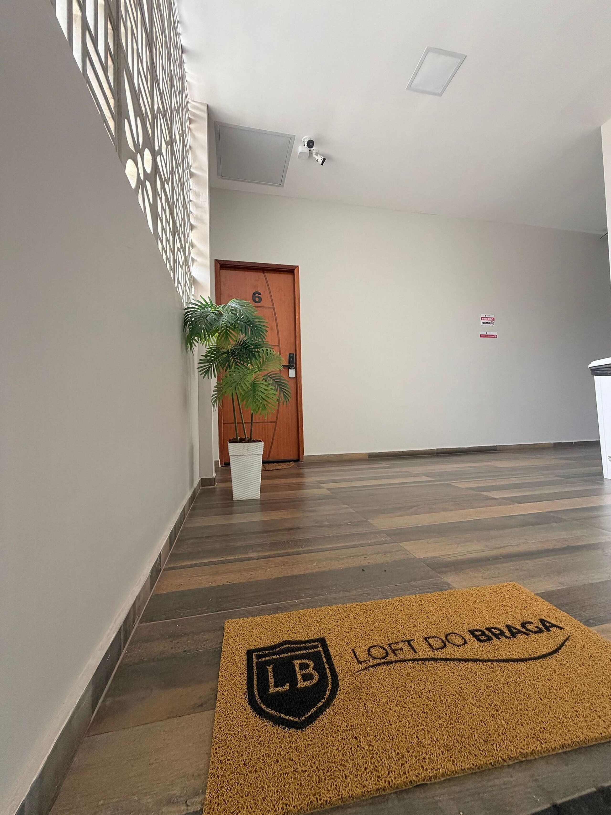 A hallway with a wooden door marked '6', a potted plant, a doormat with 'LOFT DO BRAGA' text, and a security camera on the ceiling.