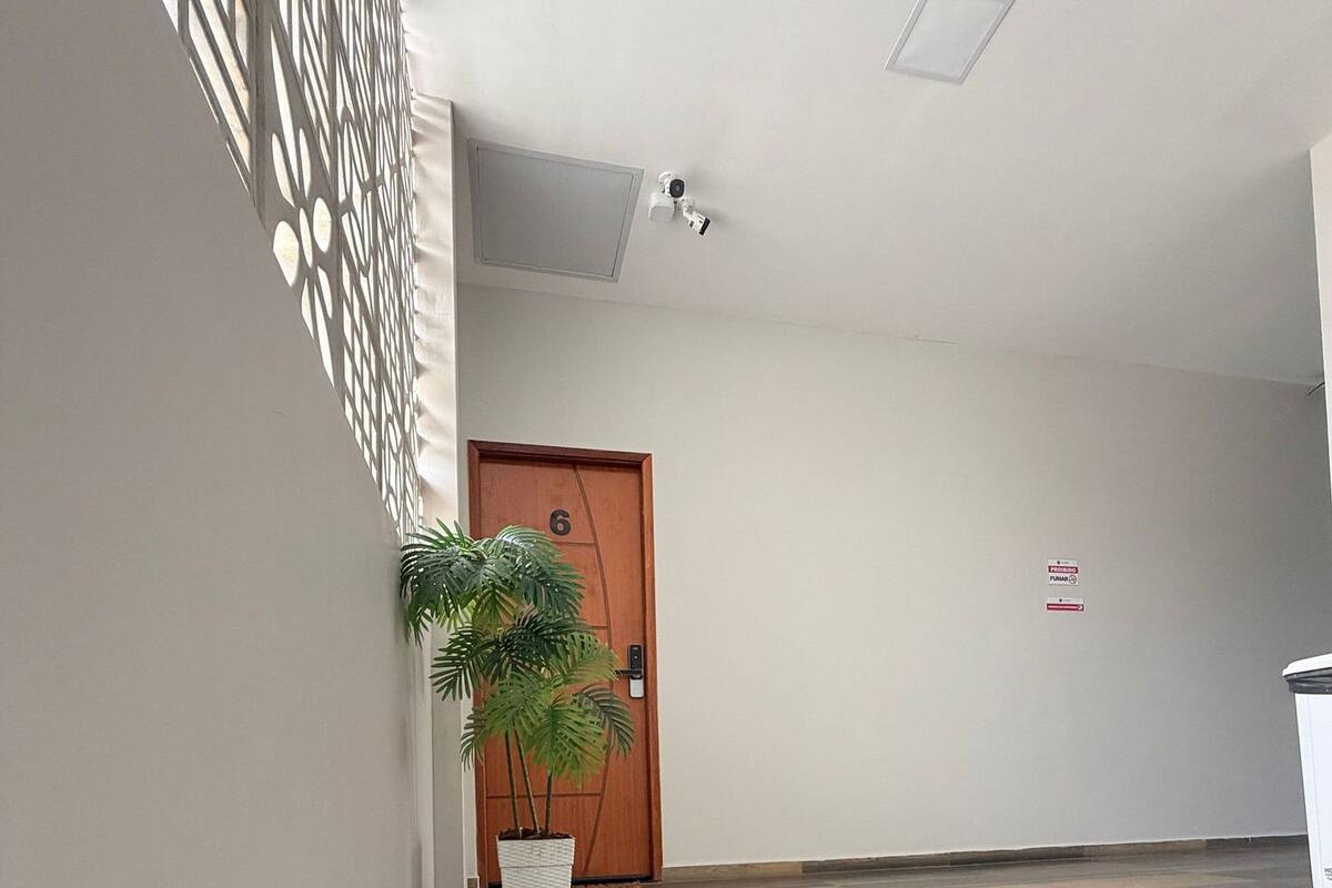 Un pasillo con una puerta de madera con el número '6', una planta en maceta, un felpudo con el texto 'LOFT DO BRAGA' y una cámara de seguridad en el techo.