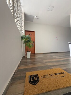 A hallway with a wooden door marked '6', a potted plant, a doormat with 'LOFT DO BRAGA' text, and a security camera on the ceiling. - Loft do Braga (Cabo Frio)