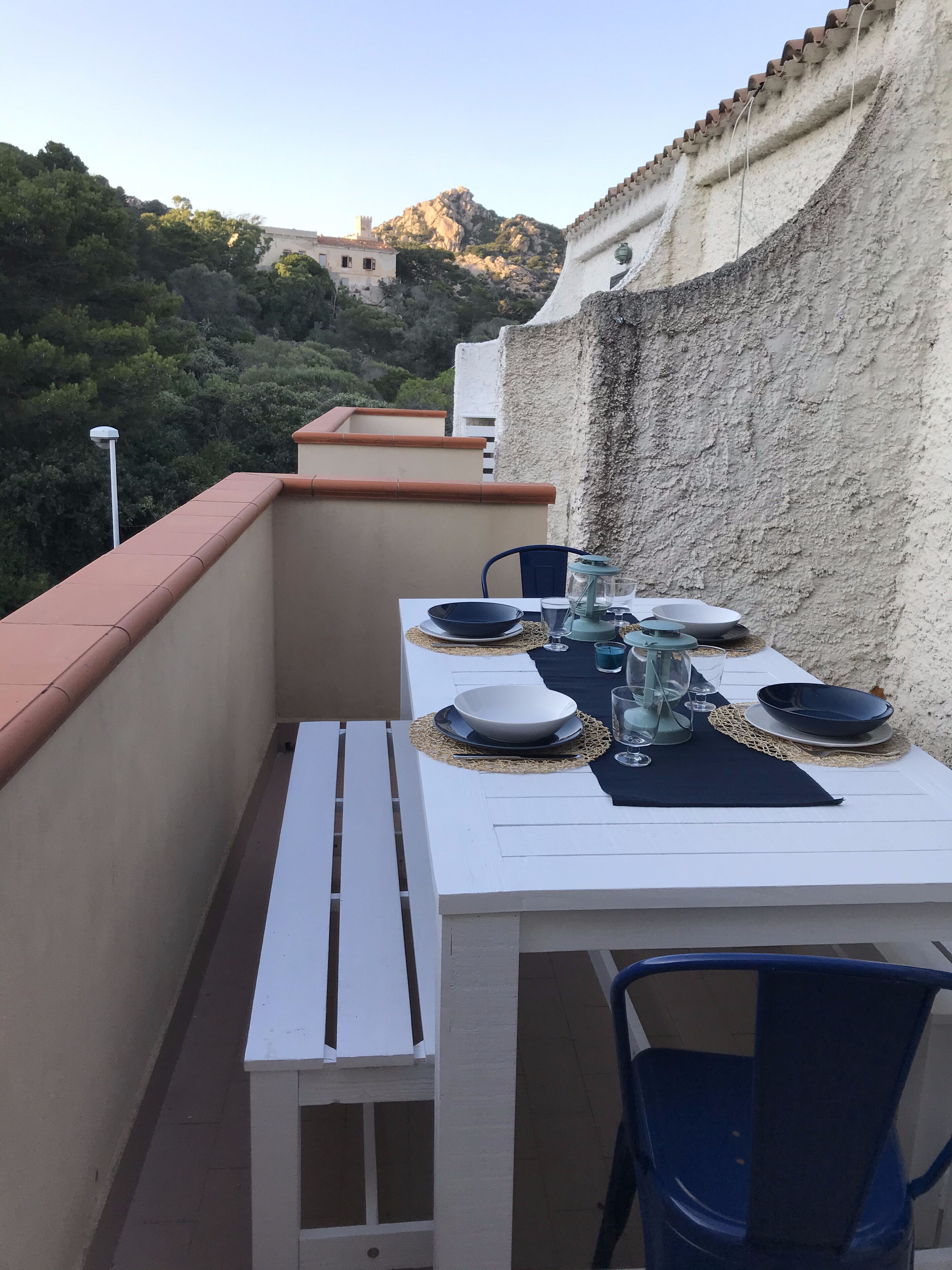 A balcony with a white table set for dining, featuring plates, glasses, and a lantern.