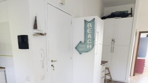 Une pièce avec un plancher en bois, des murs blancs, une porte avec un trou de serrure, une petite table avec une chaise, et un panneau indiquant : « LA PLAGE, C’EST PAR LÀ ».