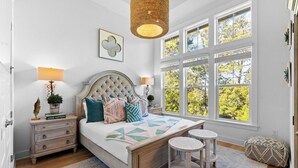 A bedroom with a large bed, two bedside tables, a hanging light fixture, and a large window with a view of trees. - Playful & Fun at Coco's~ Golf Cart, Bikes, 2 Pools PLUS Easy beach access (Inlet Beach)