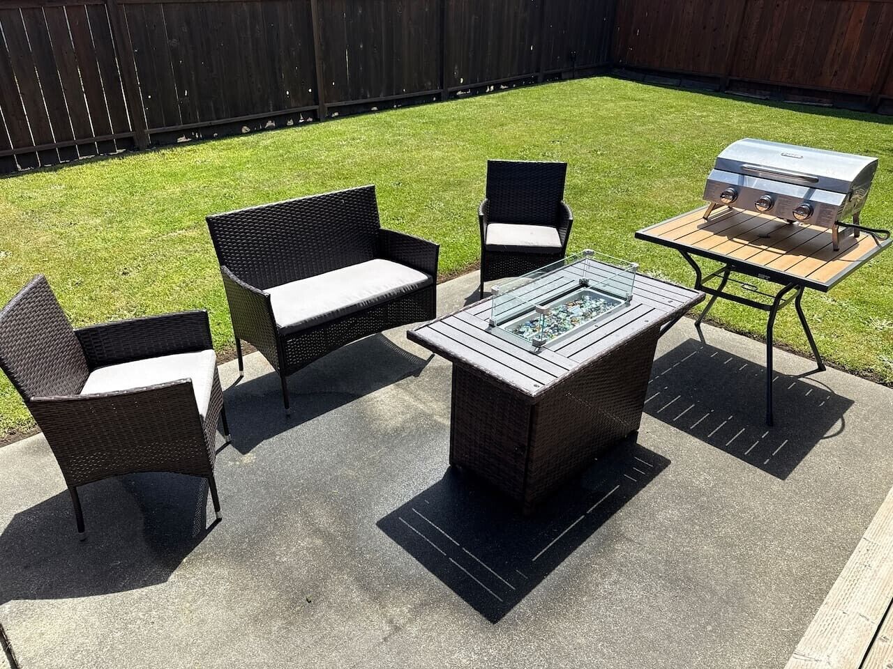 Een terras buiten met rieten meubels, een tafel met glazen blad en een barbecue.