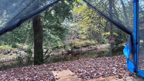 A tent set up in a forested area with a wooden deck and a sign warning about swimming risks. - Riverside cabin in the woods (Bracebridge)