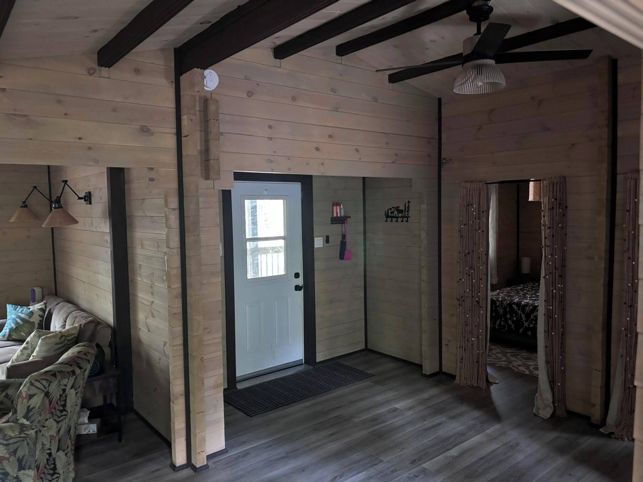 A room with wooden walls and a sloped ceiling, a bed with a patterned bedspread, a door with a window, and a wall-mounted shelf with items.
