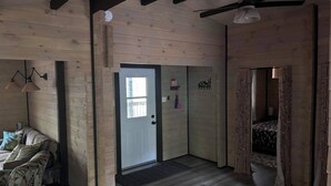 A room with wooden walls and a sloped ceiling, a bed with a patterned bedspread, a door with a window, and a wall-mounted shelf with items. - Riverside cabin in the woods (Bracebridge)