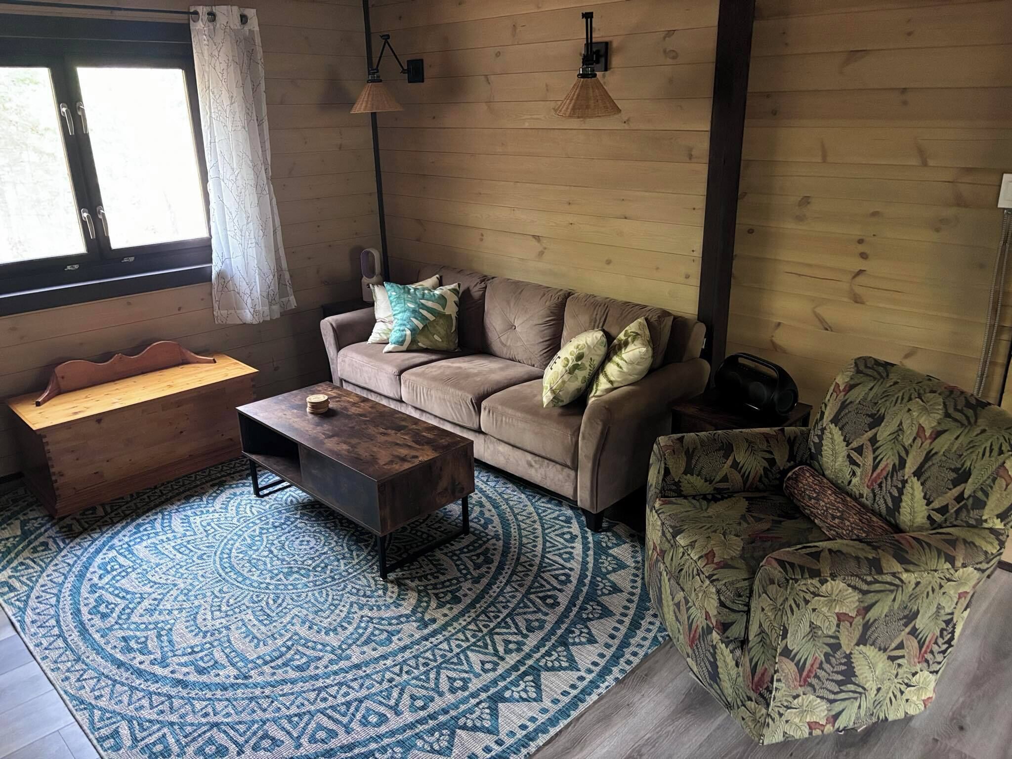 A cozy living room with a wooden floor, a patterned rug, a sofa with cushions, a bench, a coffee table, and two wall-mounted lamps.